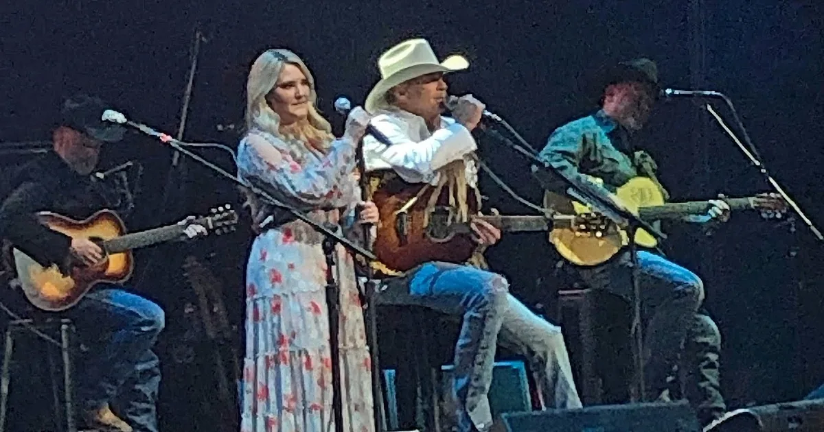 A woman in a floral dress sings on stage with a man in a cowboy hat, accompanied by musicians.