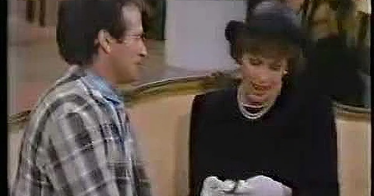 A man in a plaid shirt and another in a suit are both laughing, with the man in the suit holding a white cloth.