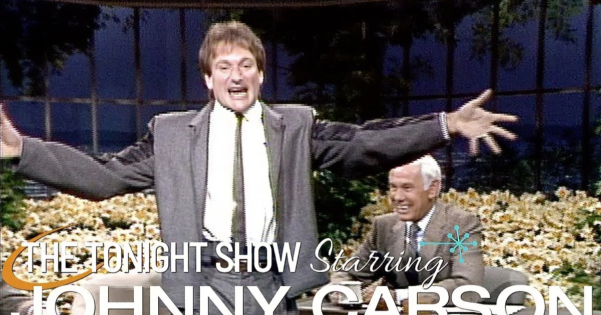 Two men in suits are seated, one laughing, the other gesturing, on a talk show set with a backdrop of flowers.