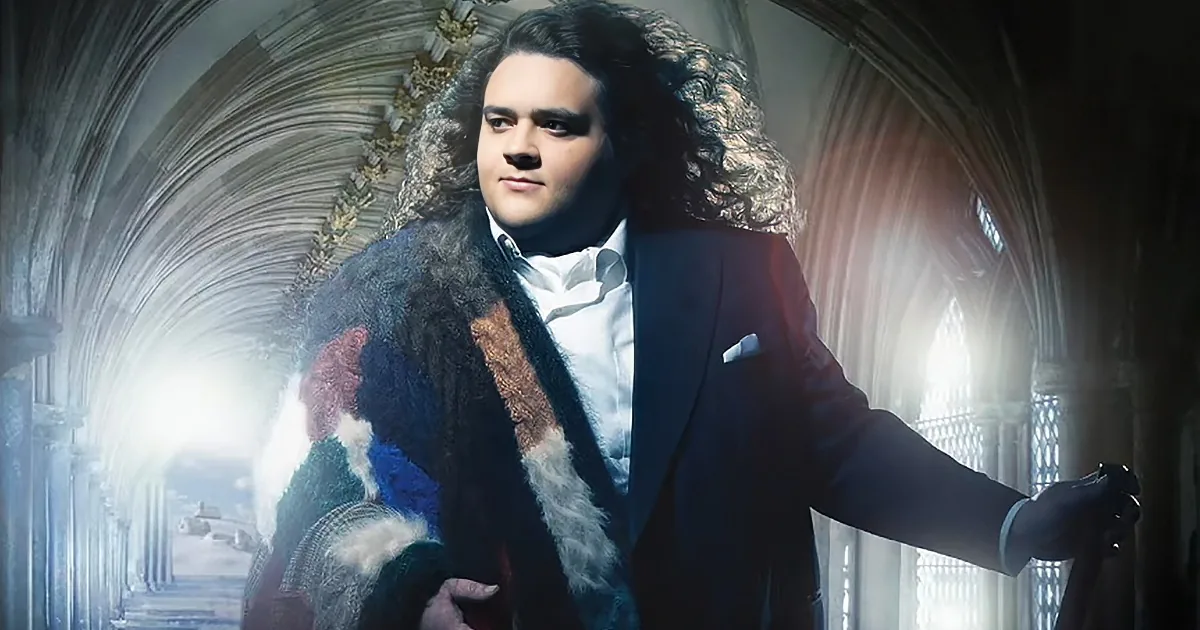 A man in a suit and fur coat stands in a grand, arched hallway with sunlight streaming through.