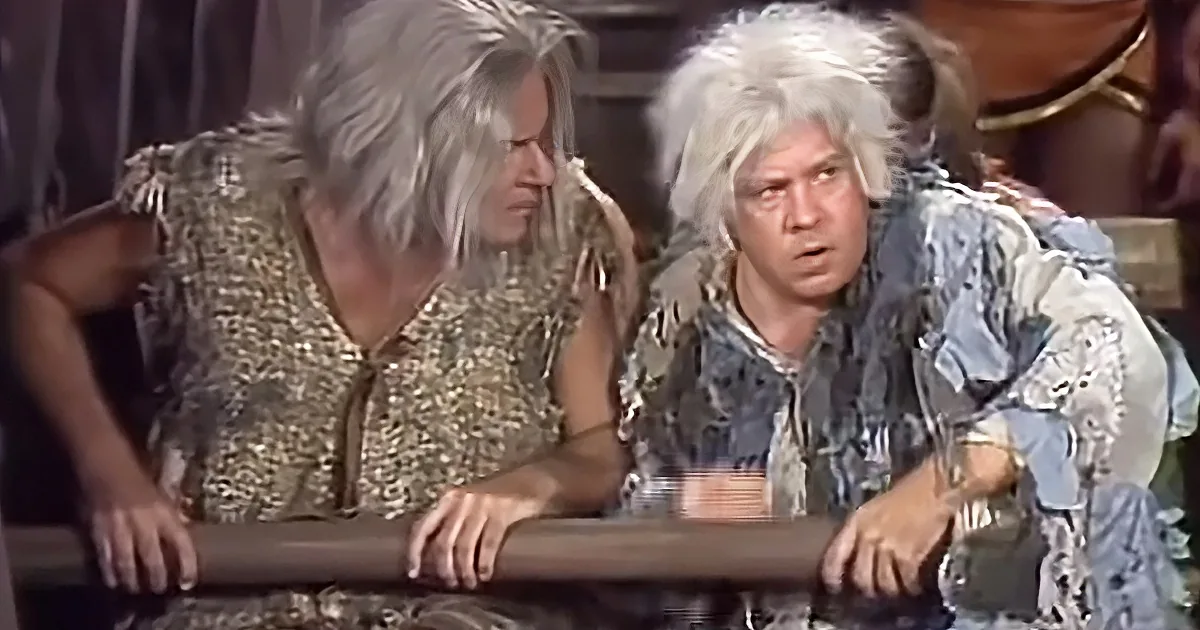Two individuals with spiky white hair, one in a gold outfit and the other in a blue outfit, are leaning over a table.