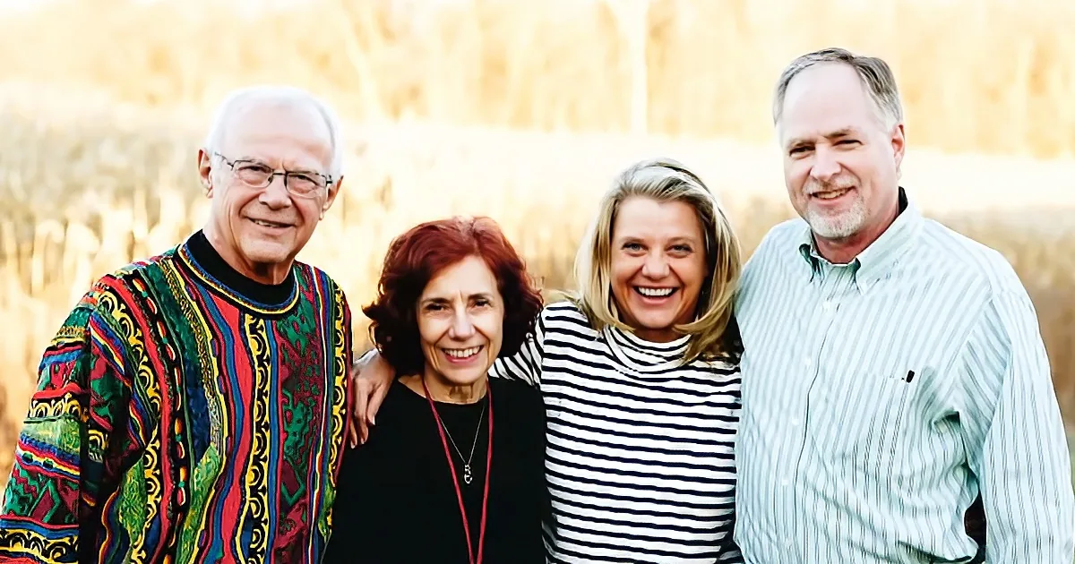 Four smiling individuals in a field, two men and two women, dressed casually, posing together.