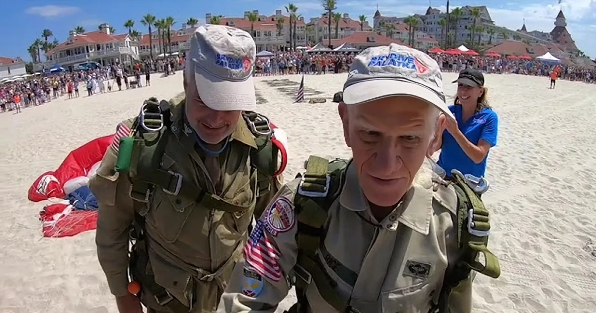 An older man in military gear is about to parachute, with a parachute visible in the sky above a coastal town.
