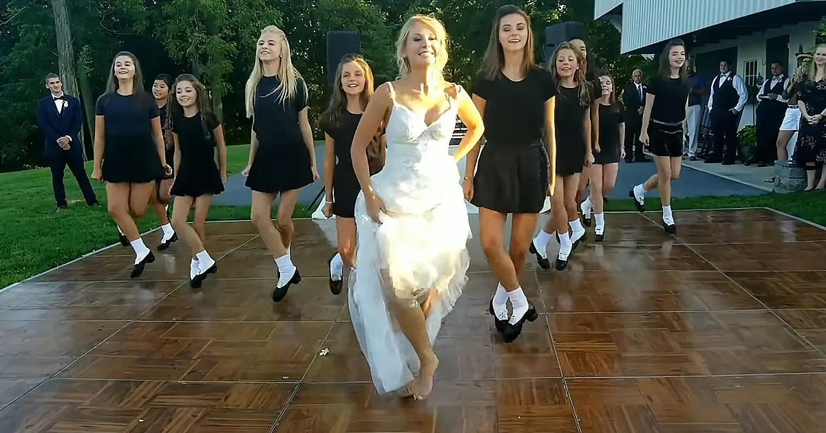A bride leads a group of dancers in a lively Irish step dance on a wooden floor.