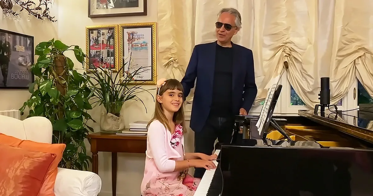 A young girl in pink plays piano, accompanied by a man in sunglasses standing nearby.