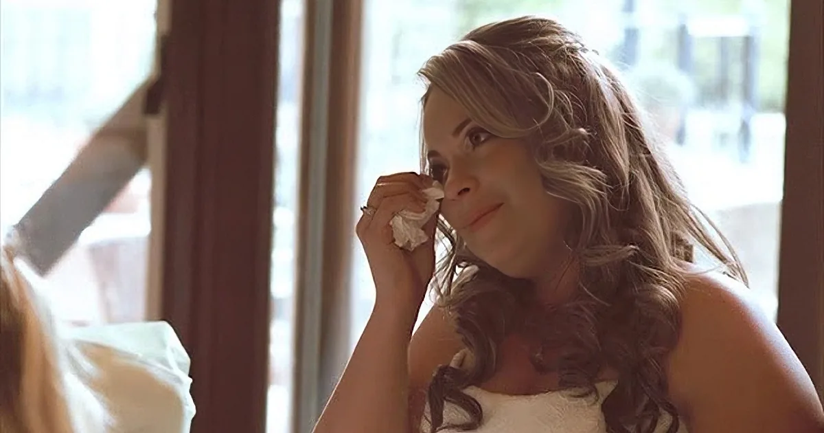 A man in a suit holding a microphone, smiling, with a blurred background of people and light.