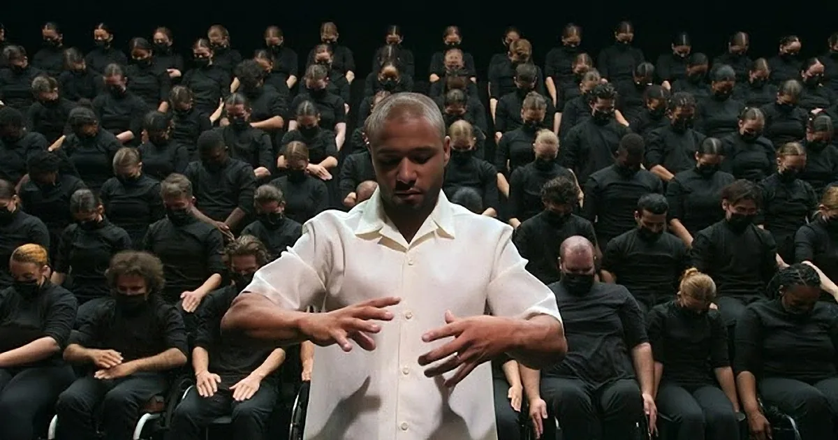 A man in white stands in front of a group of people in black, all with hands crossed in front of their faces.