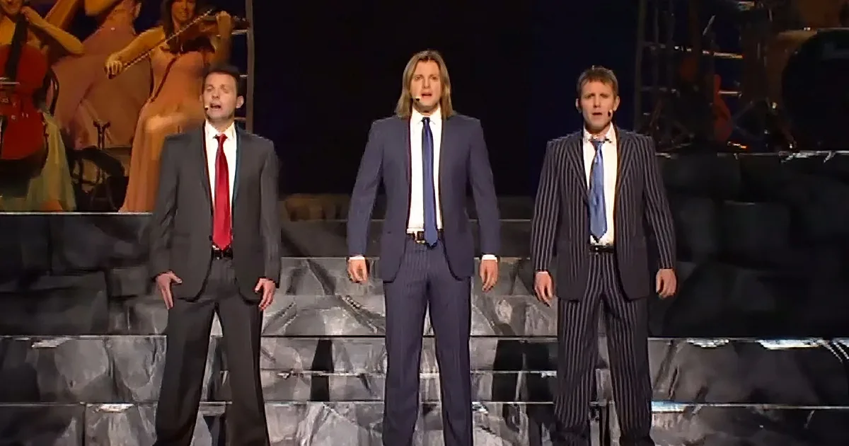 Three men in suits sing on stage with microphones, accompanied by a blurred orchestra backdrop.