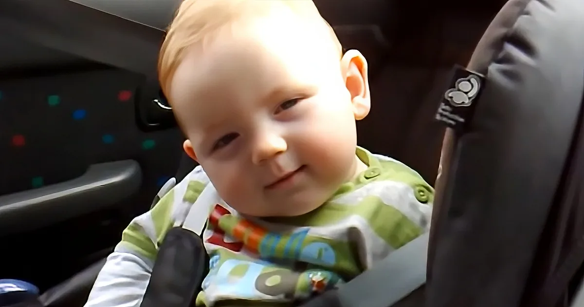 A smiling baby in a car seat, wearing a green and white shirt, with a seatbelt visible.