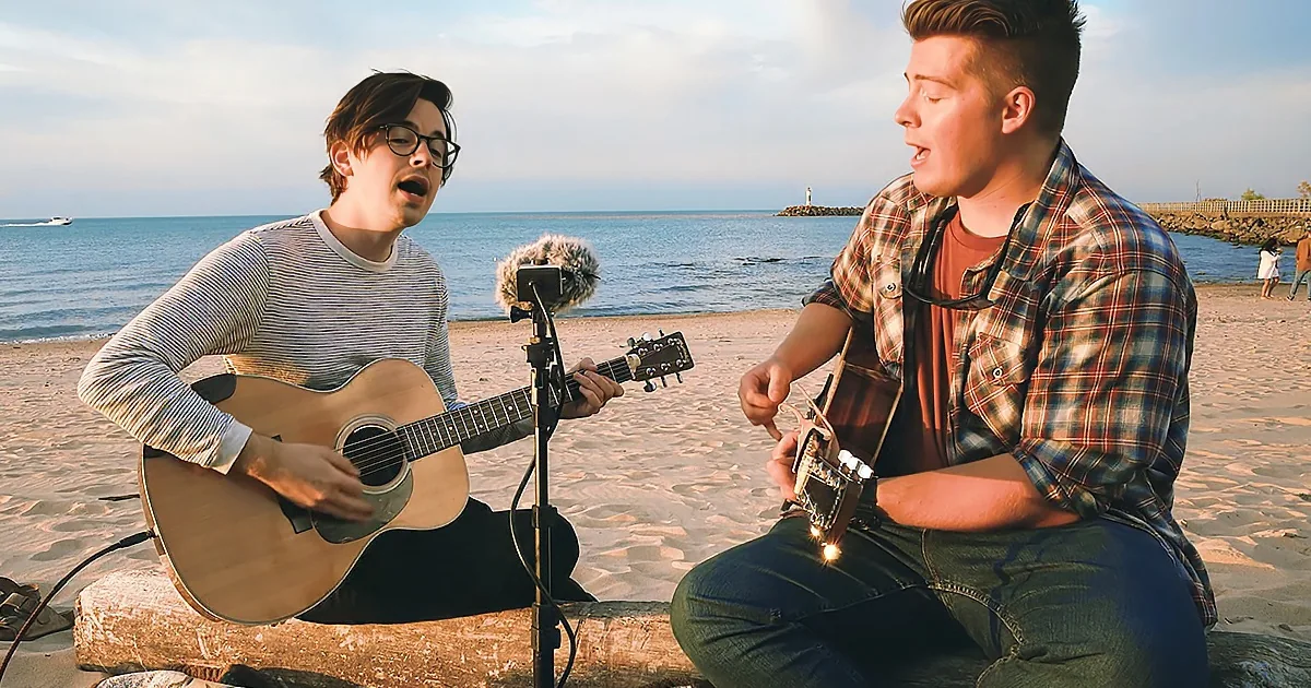 Two individuals play acoustic guitars on a beach, singing with a microphone and scenic ocean backdrop.