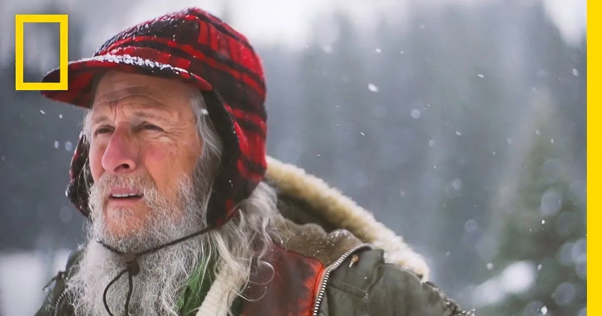 A man in a red plaid hat and winter coat smiles amidst falling snow, surrounded by trees.