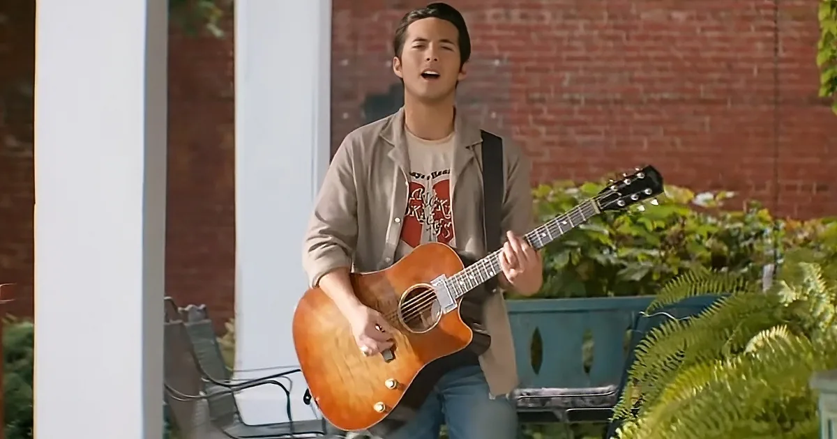 A man in a beige jacket plays an acoustic guitar outdoors, surrounded by greenery and a brick wall.
