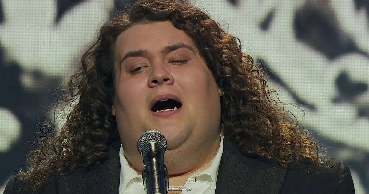 A man with long curly hair, wearing a black suit and white shirt, stands on stage with a microphone, gesturing with his...