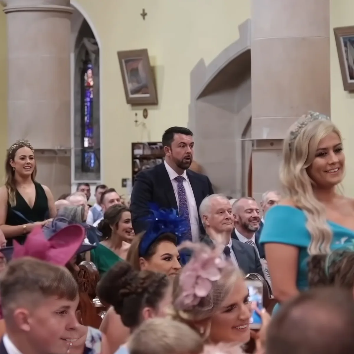 A man in a suit and tie stands in a church, surrounded by people in formal attire, likely at a wedding.