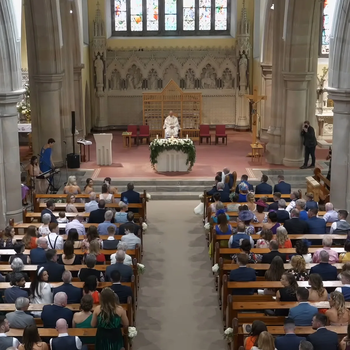 A wedding ceremony is taking place in a church with a priest at the altar and guests seated in pews.