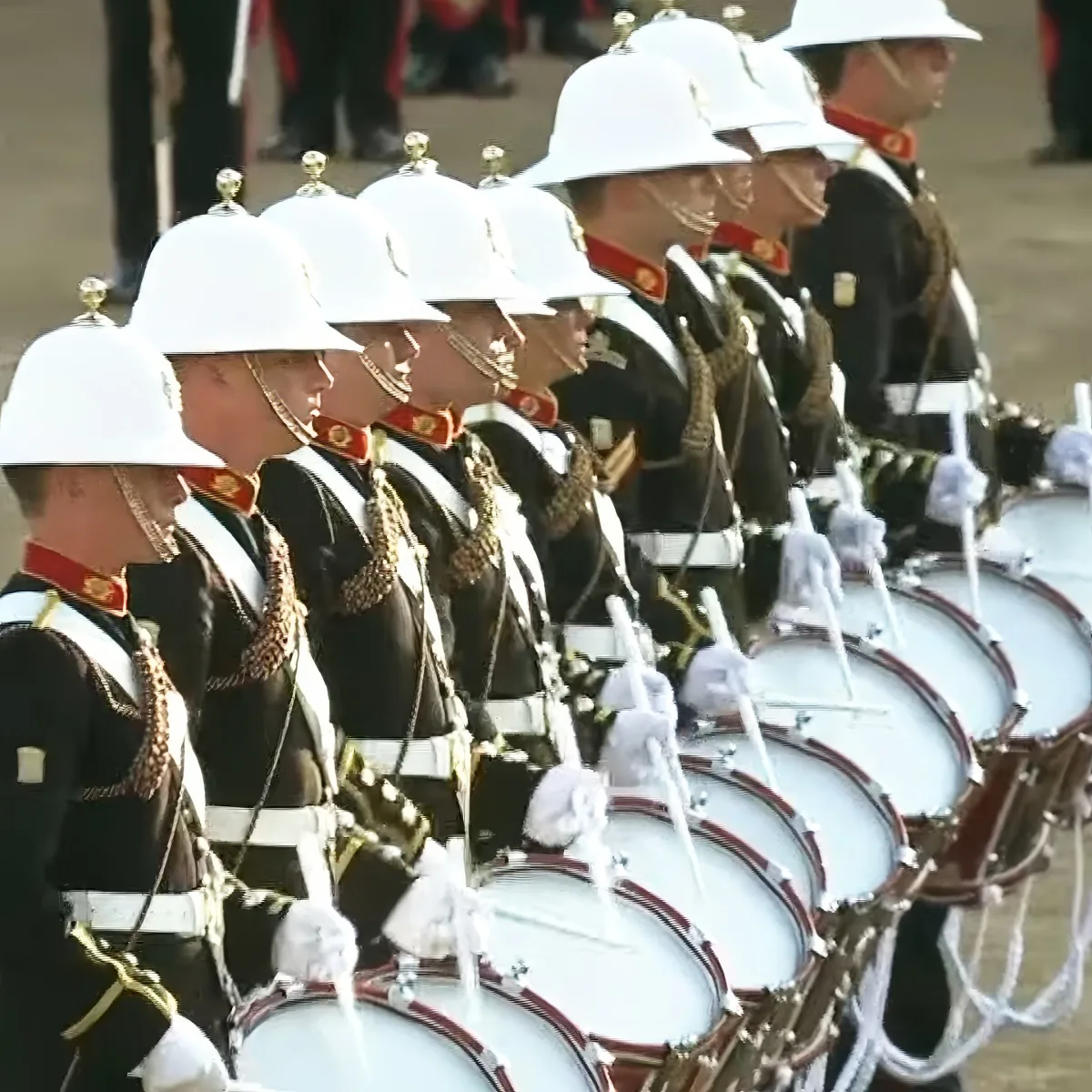 A line of uniformed drummers in white helmets and black uniforms with red accents, playing drums in a parade.
