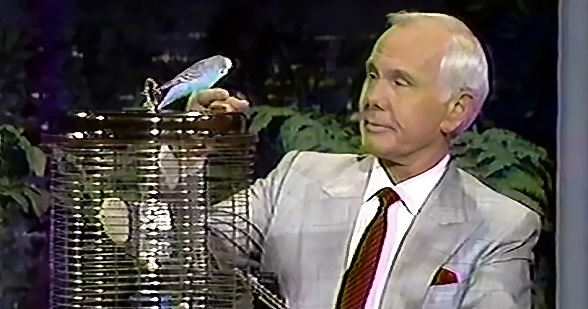A man in a suit sits beside a birdcage with a bird perched on top.