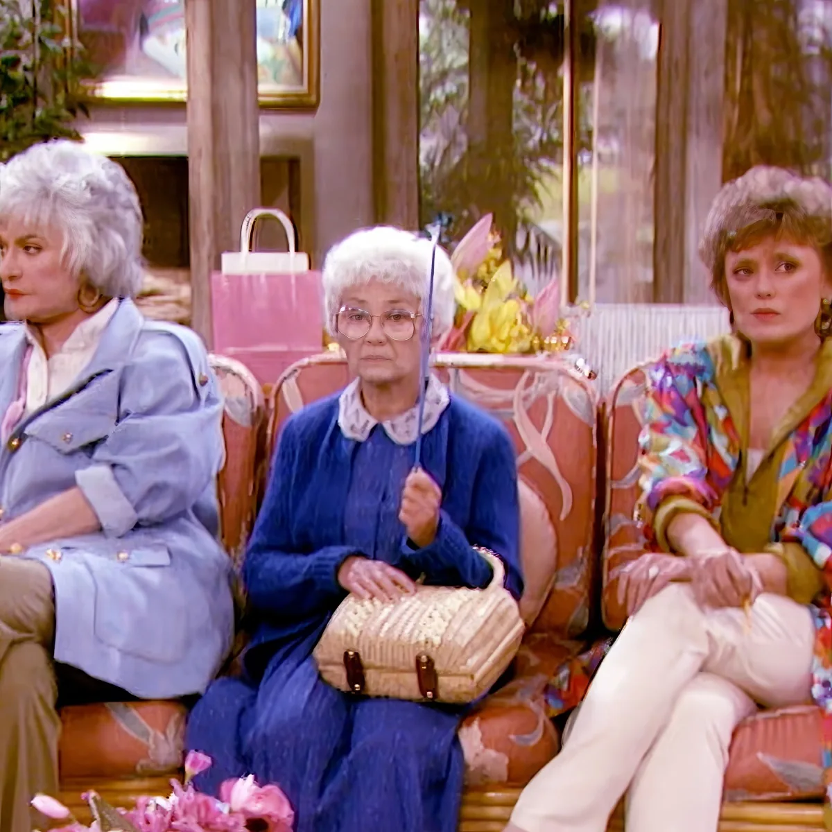 An elderly woman in a blue sweater holds a small umbrella and a beige purse while sitting on a couch.