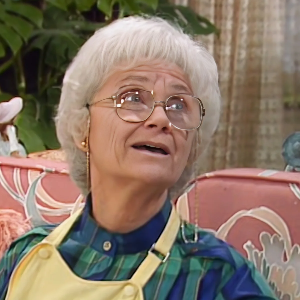 An elderly woman with white hair and glasses, wearing a green and yellow striped shirt and apron, is seated on a...
