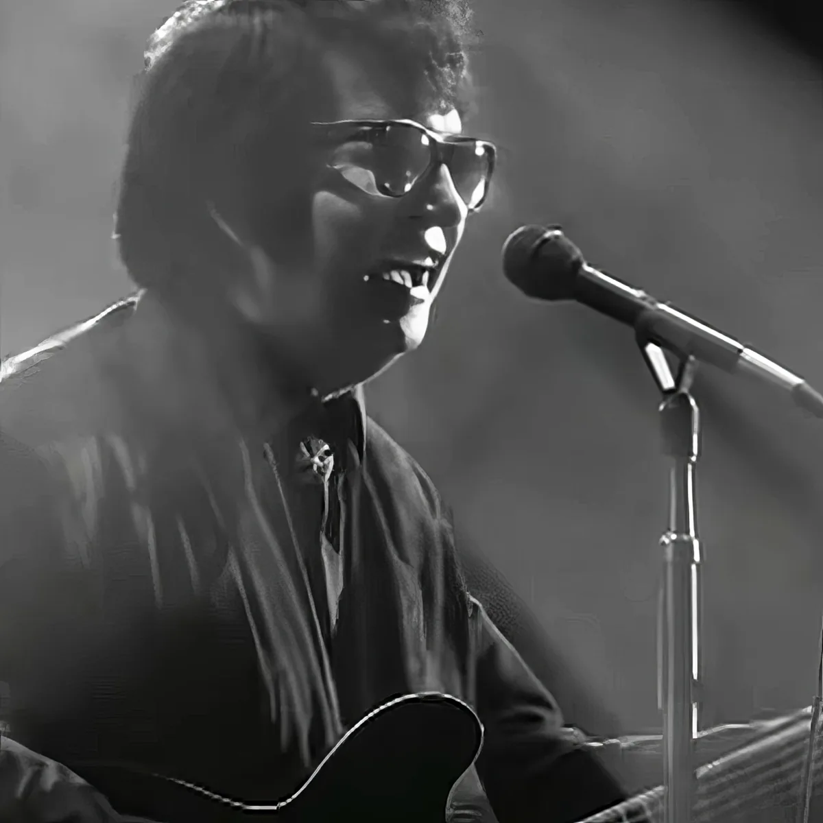 A person wearing glasses sings into a microphone while playing an acoustic guitar.
