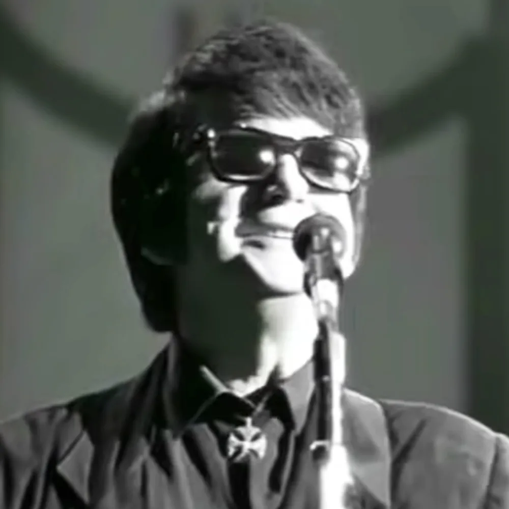 A person wearing glasses and a suit is singing into a microphone, with a dark background.