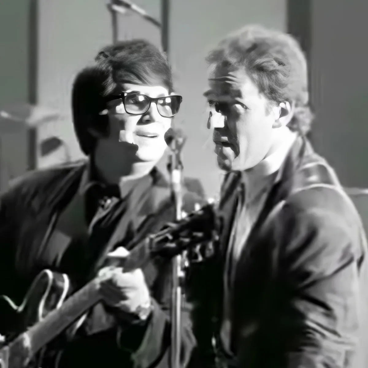 Two individuals are singing into microphones, one playing guitar, in a black-and-white setting.