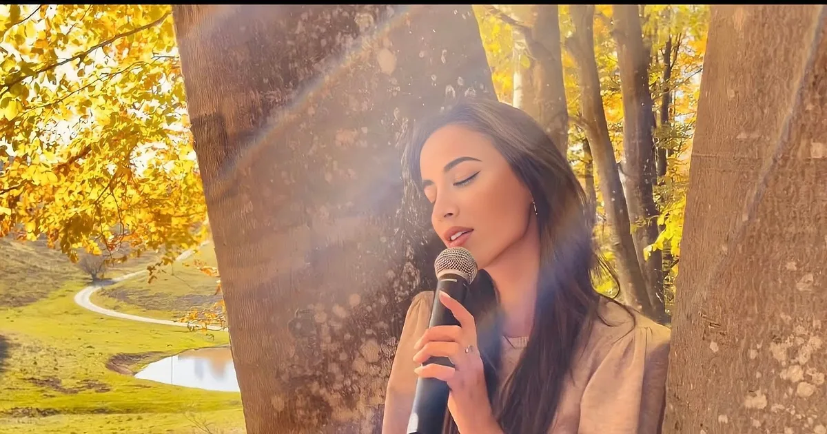 A woman sings into a microphone, surrounded by autumn trees and sunlight.