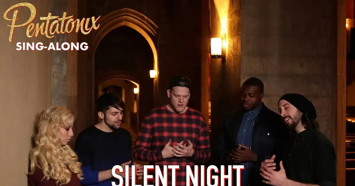 A man in a red plaid shirt stands with hands clasped, surrounded by others in a dimly lit hallway.