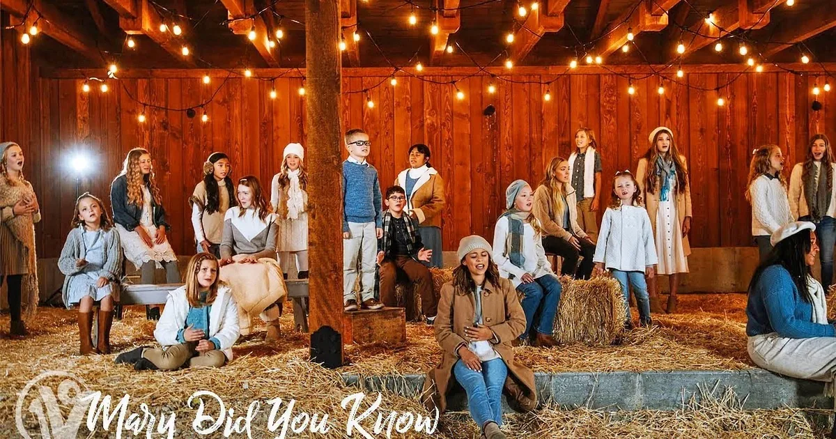 A man in a beige sweater stands in a barn with a group of people singing.