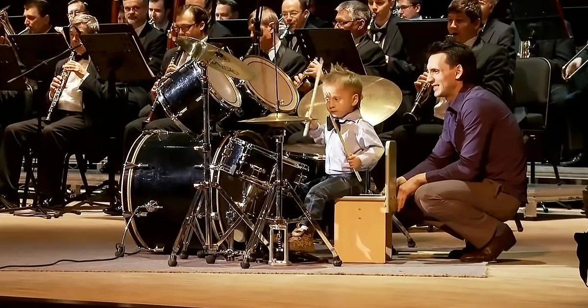 A young boy plays drums on stage with an orchestra in the background.