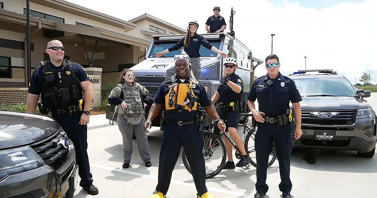 A police officer in a yellow life vest stands in front of a group of uniformed officers and vehicles.
