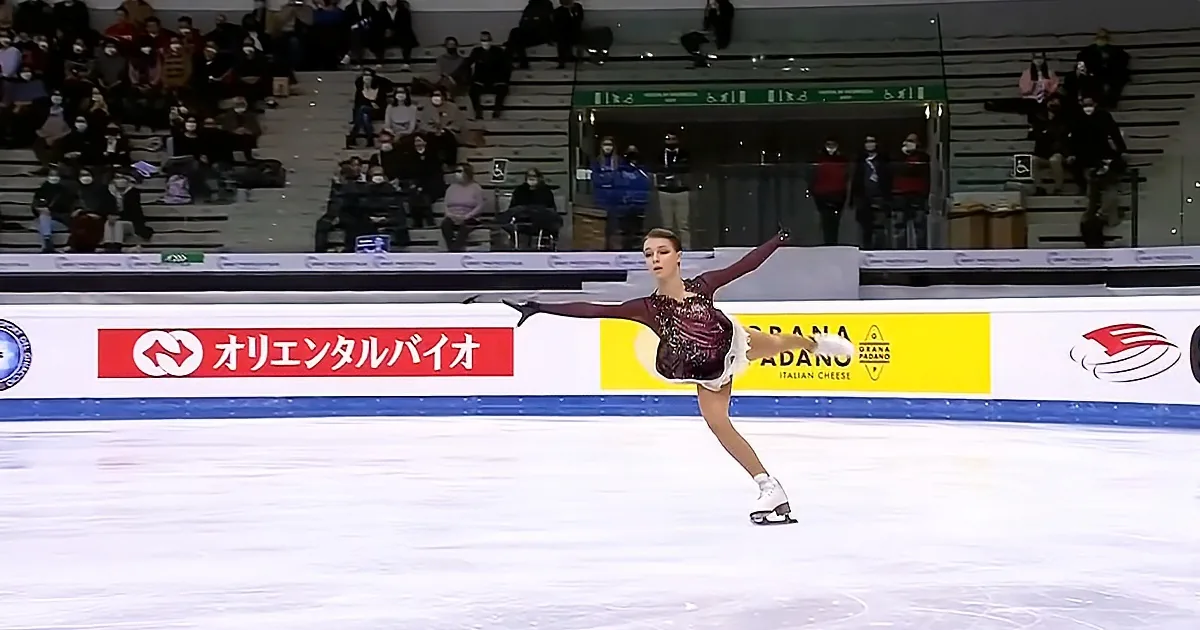 A figure skater in a maroon outfit performs a spin on the ice, with spectators in the background.