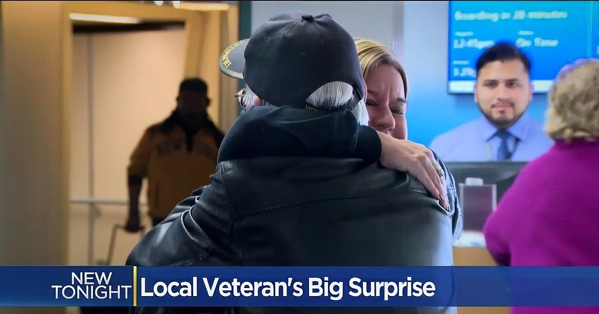 A person in a black jacket hugs another in a black beanie, surrounded by airport staff and travelers.