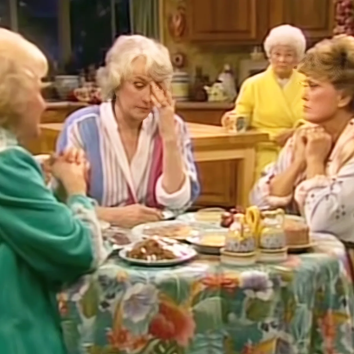 A woman in a blue robe sits at a floral tablecloth, holding her head, while others listen in a kitchen setting.