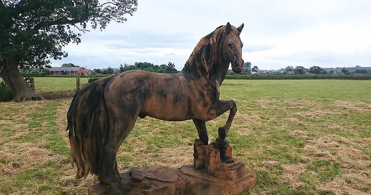 A wooden horse statue stands on a stump in a grassy field under a cloudy sky.