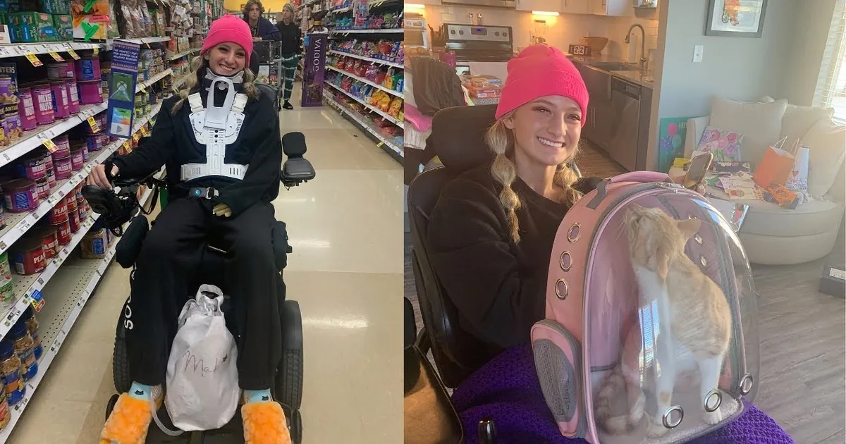 A woman in a pink beanie and black sweatshirt smiles while holding an orange cat in a cluttered kitchen.