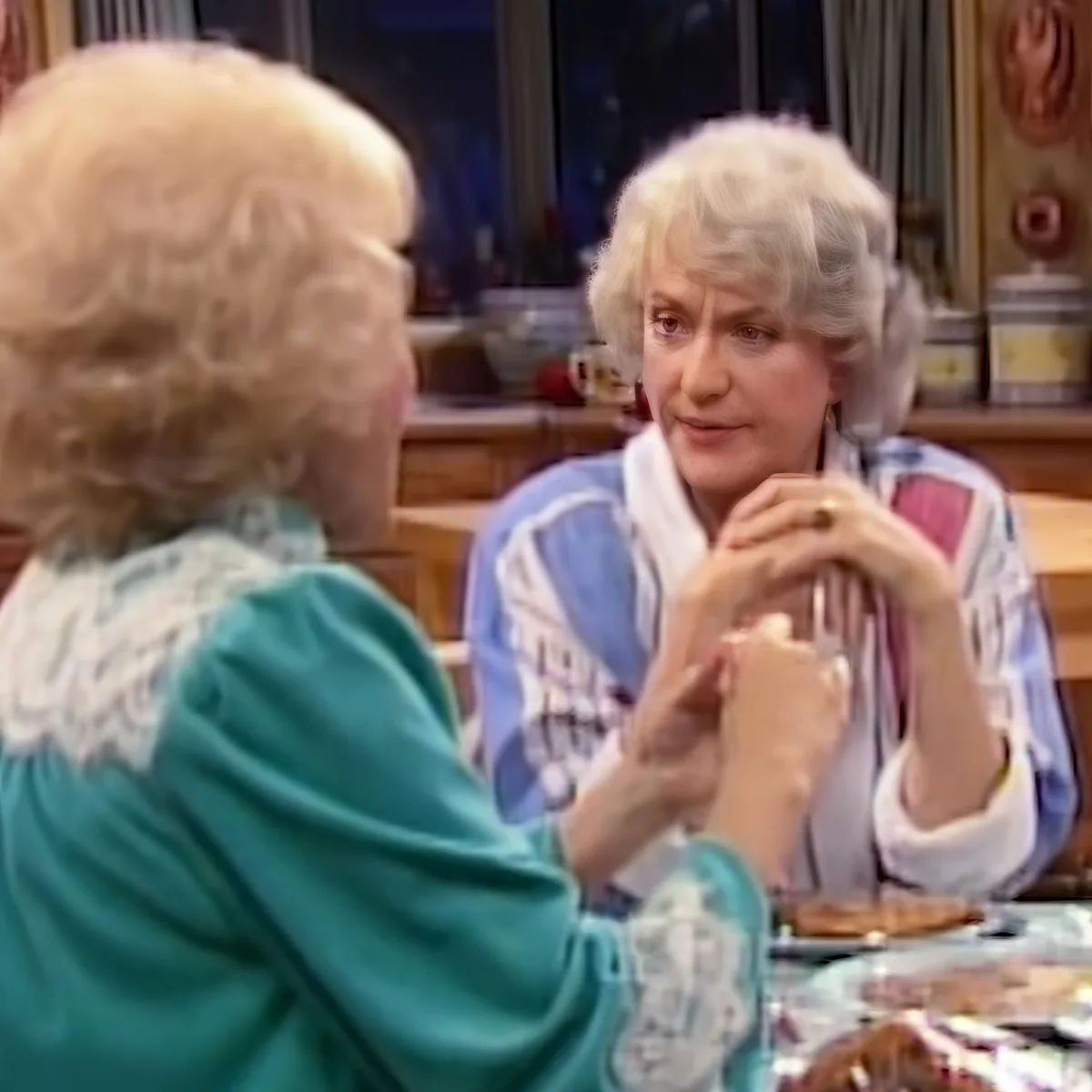 Two women are seated at a table, engaged in conversation, with one holding a fork and the other gesturing with her hands.