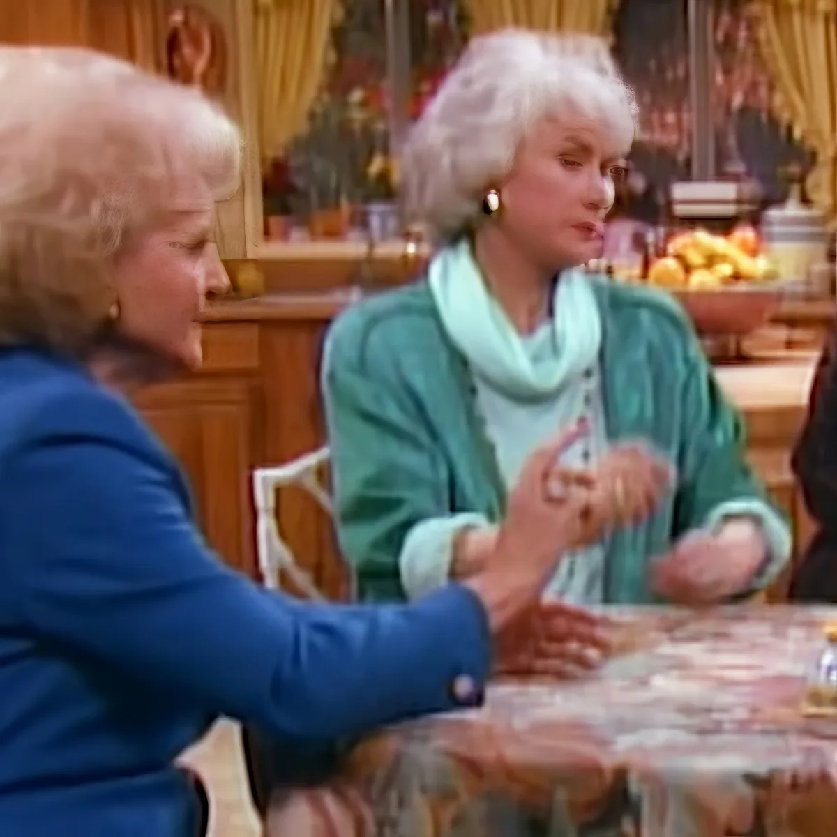 Two women are seated at a table, engaged in conversation, with a bowl of fruit and a Christmas tree in the background.
