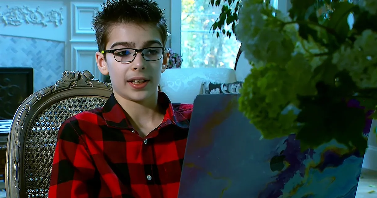 A boy in a red plaid shirt sits in a chair, holding a colorful painting, with a plant and window in the background.