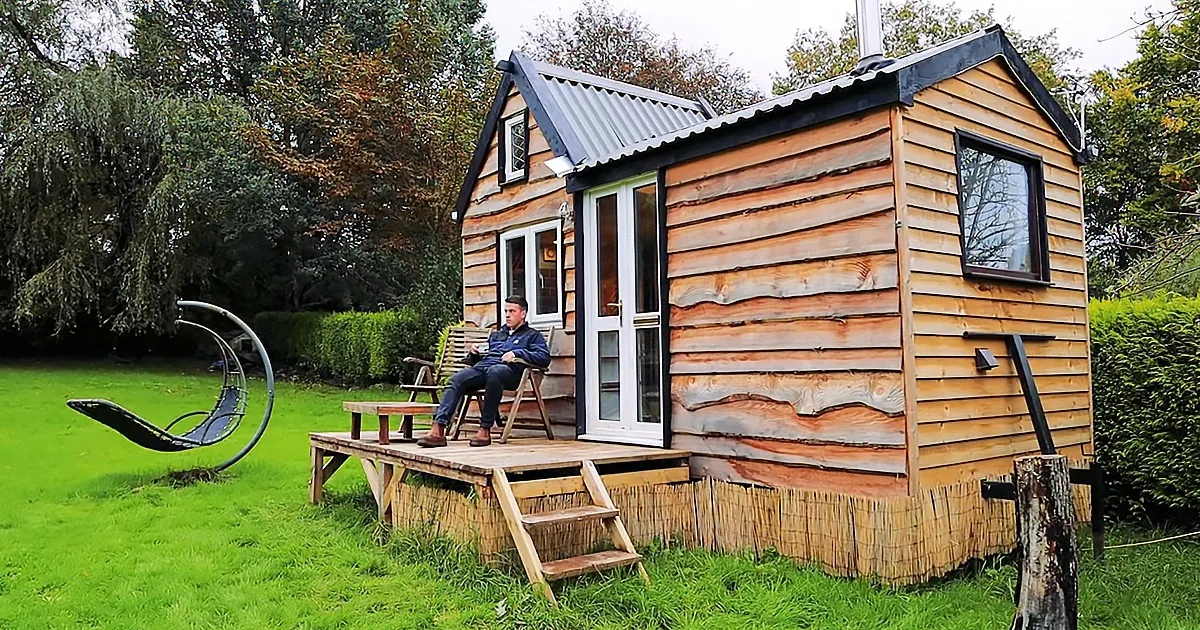 A man sits on a chair outside a small wooden cabin on a grassy lawn.
