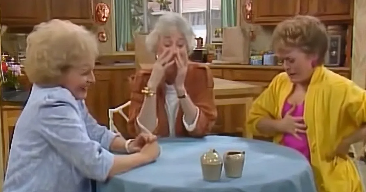 Three women in a kitchen, one covering face, another laughing, third holding chest, all around a blue table with sugar...