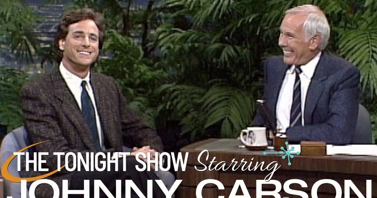 Two men in suits are seated, one laughing, in a talk show setting with a desk and plants in the background.