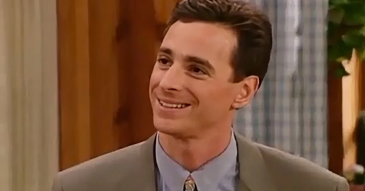 A man in a suit and tie smiles warmly, with a checkered curtain and wooden door in the background.