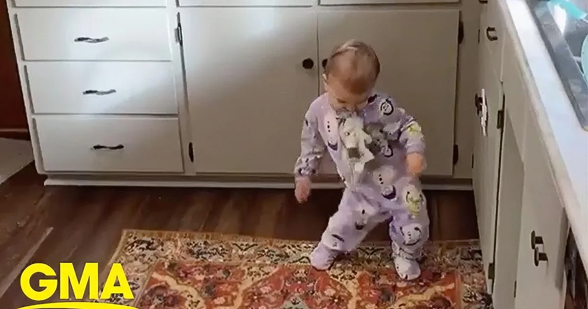 A baby in purple pajamas stands in front of a cabinet, holding a stuffed animal and looking at the camera.