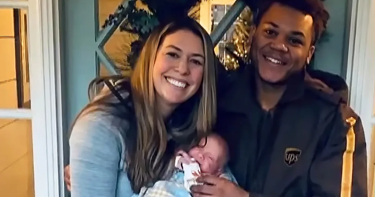 A smiling couple holds a newborn baby, standing in front of a door with a Christmas tree in the background.