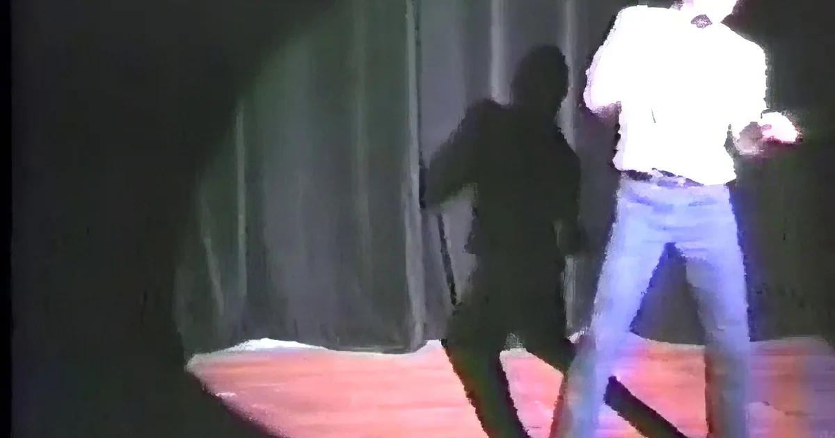 A person in a white shirt and blue pants is bent over, possibly dancing or performing a move, in a dimly lit environment.