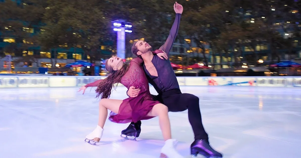 A figure skating couple performs a lift on an ice rink, with the woman in a purple dress and the man in black.