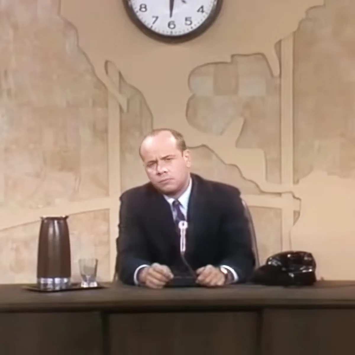 A man in a suit sits at a desk with a clock above him, appearing to be in a formal setting.
