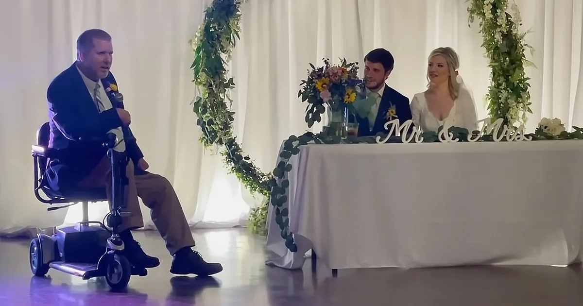 A man in a wheelchair speaks into a microphone at a wedding, with a couple seated at a decorated table.