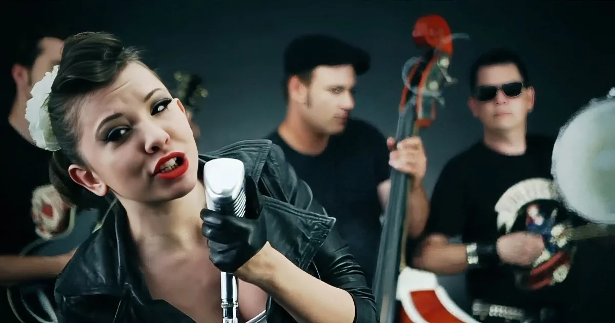 A woman in a black leather jacket sings into a microphone, surrounded by a band in a dark setting.